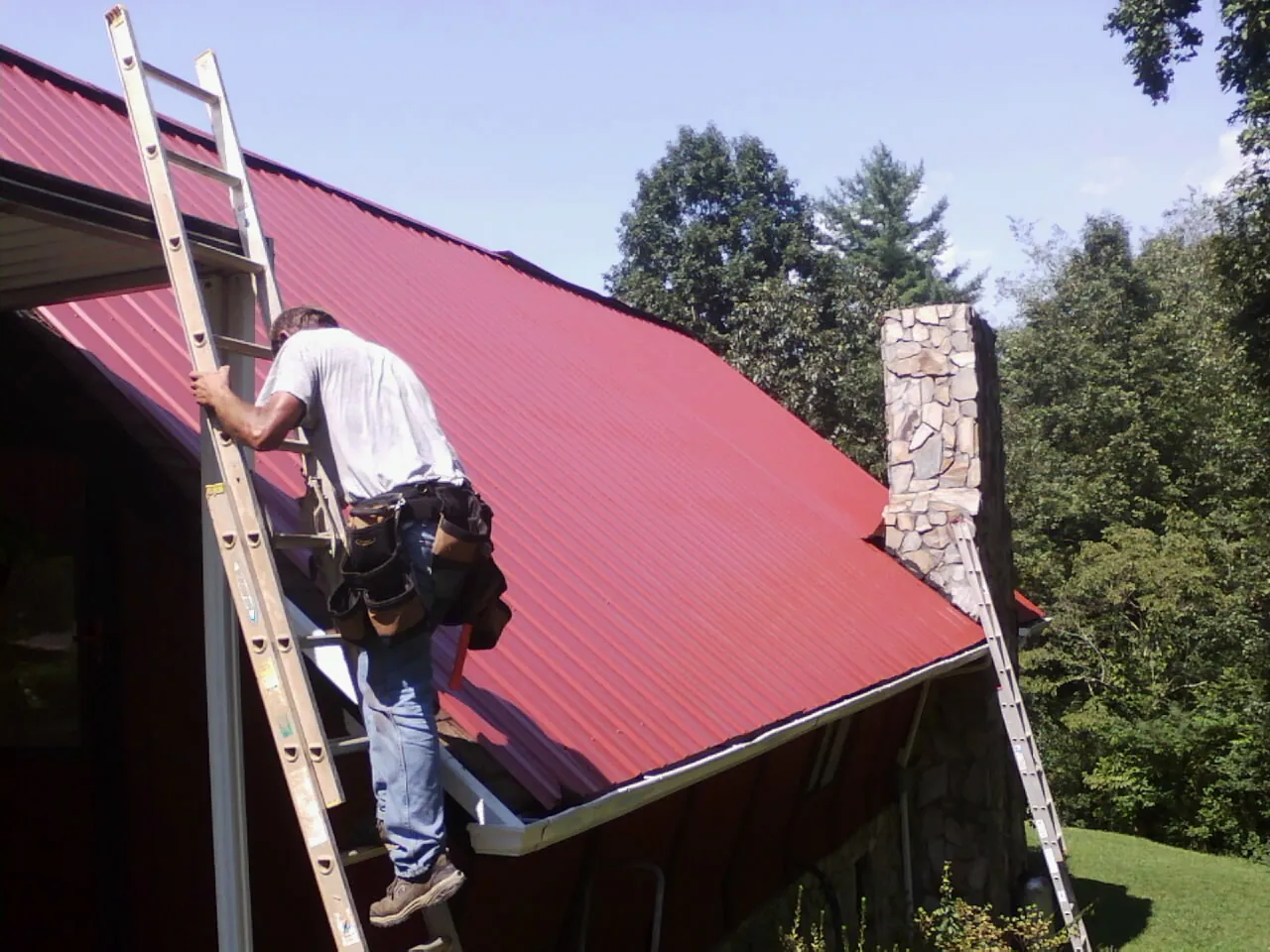 Roofer on a ladder beside a red metal roof and stone chimney — Soffit & Fascia Repair by Roofing Brothers in Cudahy